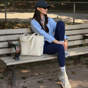 A woman sits on a wooden park bench wearing a light blue button-up shirt, navy leggings, white socks, and a black baseball cap. A cream canvas tote bag rests beside her on the bench, with trees and a chain-link fence in the background.